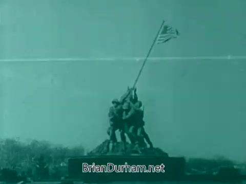 The still features a monochromatic image of the iconic Iwo Jima Memorial, depicting U.S. Marines raising the American flag. The scene is set against a muted background, emphasizing the sculpture's significance. The overall tone is somber and reverent, aligning with the theme of honoring veterans, typical of a 1960s Veterans Day PSA.