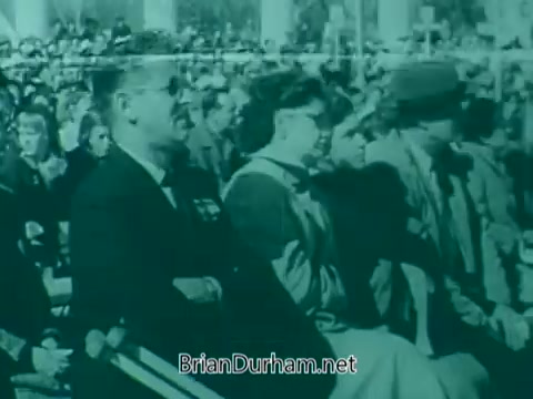 The still from the 'Veterans Day PSA - 1960's' depicts a large audience gathered for a commemorative event. Prominent figures are seated in the foreground, with a man wearing sunglasses and a woman in a light-colored outfit. The crowd appears attentive, set against a backdrop of a public gathering, suggesting a tribute to veterans. The image is presented in a vintage 16mm visual style, indicative of the era.
