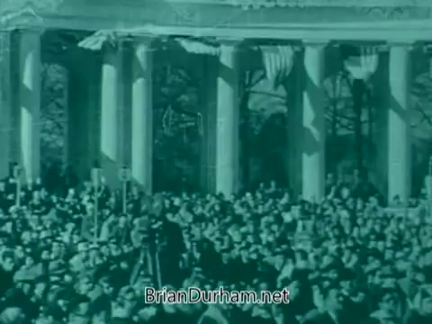 The still shows a large crowd gathered in a public space, possibly for a Veterans Day event. They are positioned in front of a classical structure marked by tall columns. The atmosphere seems solemn, and the scene is tinted in a greenish hue, suggesting it may be from an older film or video. The presence of flags or banners can be inferred, enhancing the patriotic context of the gathering.