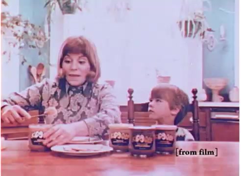 The still from the 1970s Koogle peanut butter spread commercial features a woman and a young boy sitting at a kitchen table. The woman, dressed in a patterned top, is spreading Koogle peanut butter on a plate while the boy watches attentively, showing interest in the product. There are multiple jars of Koogle peanut butter spread displayed on the table, showcasing its colorful packaging. The scene is bright and homey, indicative of the family-oriented marketing style of that era. The overall vibe is nostalgic, evoking the simplicity and charm of 1970s advertising.