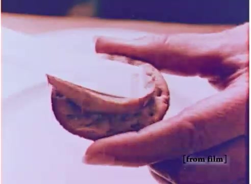 The 16mm still from the 1970s Koogle peanut butter spread commercial showcases a hand holding a small, round cookie or cracker with a generous layer of Koogle peanut butter spread on it. The spread appears creamy and is a tan or light brown color, emphasizing its peanut flavor. The visual conveys a sense of indulgence and creativity, highlighting the appealing texture and taste of the product, which is presented as a fun and innovative alternative to traditional peanut butter. The context of the advertisement suggests that Koogle not only tastes like peanuts but also comes in various flavors, making it a unique product for consumers.