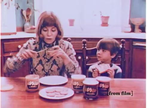 The still from the 1970s Koogle peanut butter spread advertisement features a woman and a young boy sitting at a kitchen table. The woman, dressed in a patterned top, is holding a knife and preparing to spread peanut butter on slices of bread placed on a plate in front of her. The boy, sitting beside her and looking intrigued, is holding a spoon and observing the process.   Several jars of Koogle peanut butter spread are visible on the table, showcasing different flavors. The kitchen setting conveys a warm, homely atmosphere typical of family-oriented advertisements from that era. The overall color palette suggests a nostalgic, vintage feel.