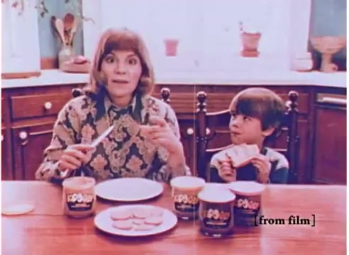 The still captures a nostalgic moment from a 1970s Koogle peanut butter spread commercial. In a cozy kitchen setting, a woman with a vintage hairstyle and patterned clothing sits at a wooden table, excitedly holding a knife, likely to spread Koogle. Next to her, a child with a curious expression holds a sandwich, emphasizing the appeal of the product. The table features jars of Koogle spread in various flavors, highlighting the brand's unique selling point of offering tastes beyond traditional peanut butter. The overall vibe is warm and inviting, typical of family-oriented advertisements from that era.
