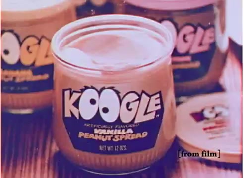 The still features a jar of Koogle peanut spread prominently in the foreground, showcasing its playful label. The jar displays the product name 'KOOGLE' in a bold, colorful font along with the flavor 'Vanilla Peanut Spread.' The background is slightly blurred, hinting at other jars of Koogle in different flavors, creating a nostalgic 1970s vibe. The overall aesthetic emphasizes the fun and unique nature of the product, capturing the essence of the era's advertising. The text suggests the spread is artificially flavored, highlighting its distinctive taste appeal.