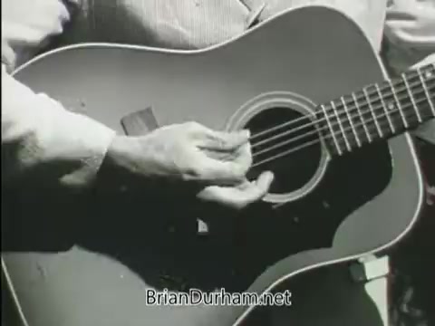 The still features a close-up of a person's hand strumming a guitar. The focus is on the instrument, highlighting its body and strings, emphasizing the act of playing music. The context suggests it may relate to themes of service or youth, typical of public service announcements from that era.