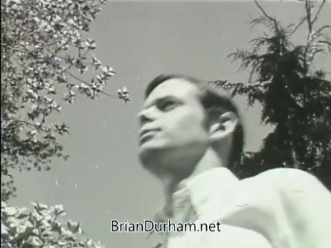 The still from the 'Junior GI Bill PSA - 1968' features a young man, likely a college student, walking outdoors. He appears thoughtful, with nature visible in the background, including trees and foliage. The black-and-white contrast emphasizes the mood and theme of the PSA, which promotes educational benefits for veterans. The overall composition suggests a sense of optimism and possibility for the future.