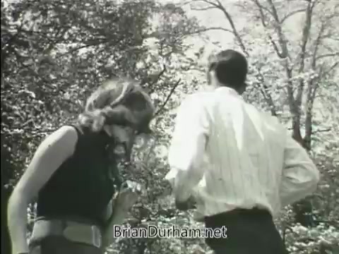 The still from the 1968 'Junior GI Bill' PSA features a young woman and a man outdoors, both dressed casually. The woman, wearing a sleeveless top and a belt, is partially turned towards the camera, while the man, in a light-colored button-up shirt, faces away. The background is filled with trees, indicating a natural setting. The scene likely emphasizes themes of youth and opportunity related to the GI Bill.