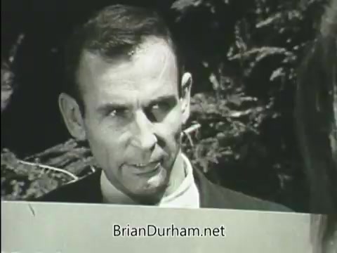 The still from the 1968 'Junior GI Bill PSA' features a middle-aged man speaking directly to the camera with a serious expression. He appears to be engaged in a discussion, likely about the benefits of the Junior GI Bill. The background includes natural elements, suggesting an outdoor setting. The focus is on his message, indicating the importance of the initiative for young veterans.
