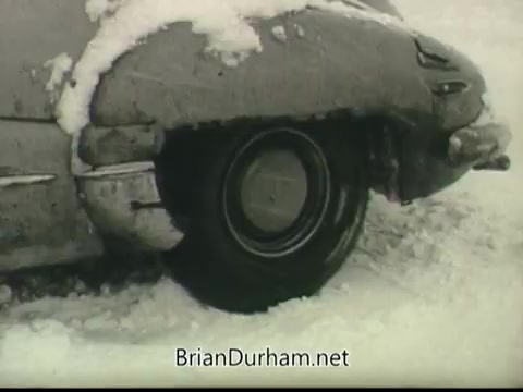 The still shows a close-up of a car partially buried in snow, with a focus on the wheel and a section of the vehicle's body. The scene conveys a harsh winter environment, emphasizing the challenges posed by severe weather conditions. The use of black and white film adds to the dramatic effect, typical of public service announcements from that era.