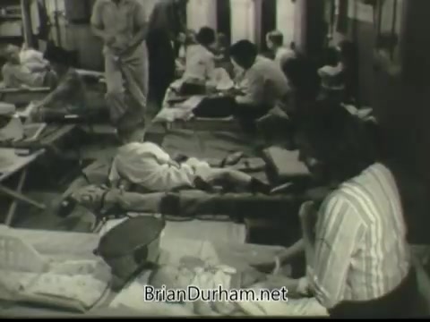 The still from the 1968 PSA 'If Disaster Strikes' depicts a crowded emergency shelter setting. Individuals, including adults and children, are shown lying on cots, suggesting a humanitarian response to a disaster. The environment appears tense yet organized, with people attending to the needs of those resting. The scene illustrates themes of community support and preparedness in crisis situations.