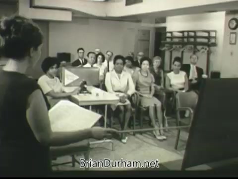 The still from the 1968 PSA 'If Disaster Strikes' depicts a classroom setting where a woman is presenting information to an audience. The audience, composed mainly of women and a few men, appears attentive while seated in chairs arranged in rows. The presenter holds a script or notes and utilizes a pointer directed towards a visual aid or a diagram on an easel, emphasizing educational content related to disaster preparedness. The overall atmosphere suggests a focus on community awareness and safety measures.