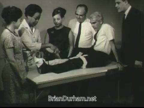 The still features a group of six individuals gathered around a table. One person, presumably a victim, is seated with an injured leg elevated and bandaged. The others are actively engaged in providing care or assessing the injury. The scene conveys a sense of urgency and teamwork, emphasizing the importance of first aid and community response in crisis situations. The overall tone is instructional, fitting for a public service announcement.