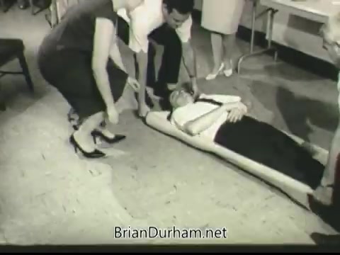 The still from the 1968 PSA 'If Disaster Strikes' shows a group of people assisting an unconscious man lying on a stretcher. Two individuals are positioned at the head and feet of the stretcher, while a woman kneels nearby observing. The setting appears to be indoors, possibly in a classroom or training environment, highlighting emergency response training and preparedness.