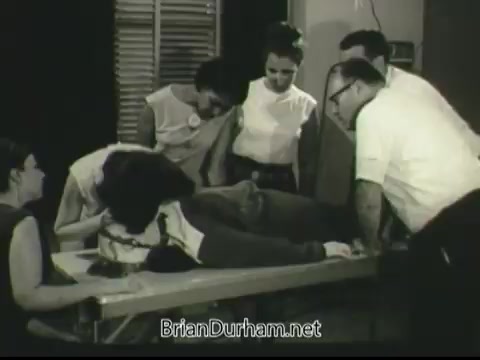 The still from the 1968 PSA 'If Disaster Strikes' depicts a group of people gathered around a patient lying on a hospital bed. The scene suggests a medical or emergency situation, with several individuals attentively observing or assisting, conveying a sense of urgency and teamwork in response to a crisis. The black-and-white format emphasizes the period's aesthetic and seriousness of the topic.