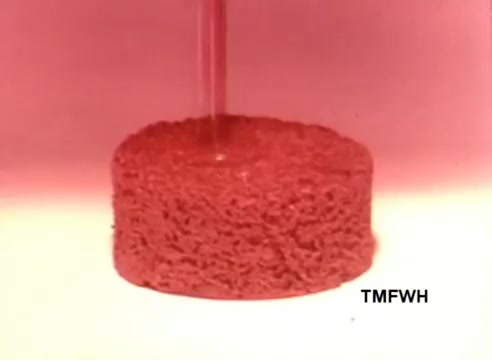 The still shows a close-up of a round, cake-like pastry, possibly a Hostess Ding Dong. A stream of a liquid, likely chocolate or icing, is being poured over it, creating a vibrant contrast against the pale background. The retro aesthetic reflects the quirky vibe of 1970s commercials, capturing a playful, indulgent moment associated with the snack. The text “TMFWH” is visible in the corner.