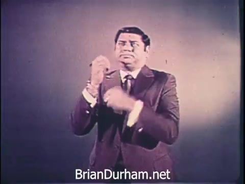 The still from the 'Niagara Rolla-ssage chair commercial' (1968) features a man in a suit, possibly mid-action, conveying a sense of urgency. The background is simple, focusing attention on the speaker. The commercial emphasizes the importance of self-care for busy individuals, suggesting that the Niagara Rolla-ssage chair offers crucial relaxation. The tone highlights the stress of decision-making and the value of taking care of oneself amidst a hectic lifestyle.