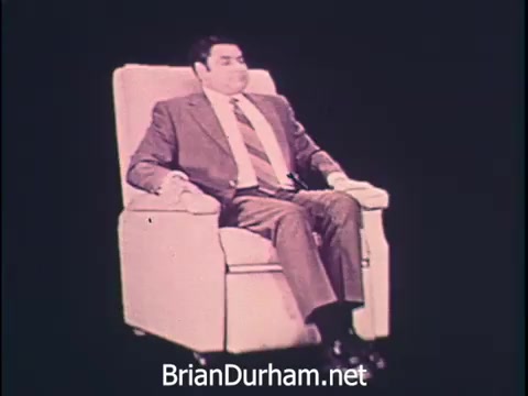The still from the 1968 Niagara Rolla-ssage chair commercial features a man seated in a comfortable, plush chair. He is dressed in business attire, indicating a professional setting. The backdrop is likely a simple, unobtrusive color to emphasize the chair's comfort. The accompanying narrative emphasizes the importance of relaxation for busy professionals, promoting the chair as a means to achieve comforting rest amidst a hectic lifestyle. The overall message is one of self-care for those who work hard.