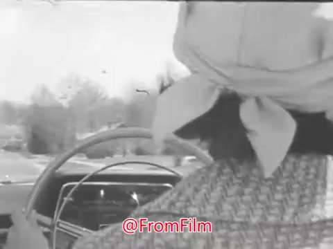 The still from the 1969 Aetna Automobile Safety PSA titled 'Inattention' features two women in a car, with one driving. The interior view shows a classic car dashboard and the driver's hands on the wheel. The women are engaged in a casual conversation about fashion and outfits. The setting appears to be outside, with trees in the background, indicating they are driving. The context suggests a lighthearted moment that contrasts the theme of the PSA, focusing on the dangers of distraction while driving.