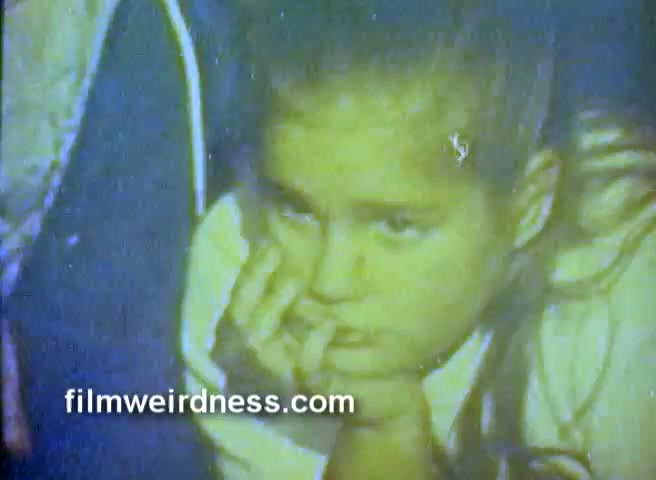 The still captures a young girl with a contemplative expression, resting her chin on her hand while looking off-screen. The colors are muted, suggesting a faded quality typical of older 16mm films. This snippet is likely from a commercial for Barney’s NY, with a nostalgic ambiance characteristic of the early 1970s. The context hints at a combination of engaging advertisements alongside a charming, possibly festive narrative.