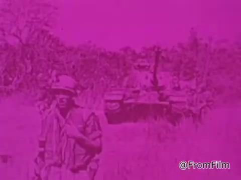 The still from the 1968 PSA 'Mail Early to Your Soldier' features a soldier in a combat uniform, set against a backdrop that appears altered with a bright pink hue, giving the image a surreal quality. In the background, an armored tank is visible, suggesting a military context, likely in Vietnam. The soldier is holding a weapon, and there is foliage surrounding the scene, typical of the Vietnam landscape. The overall tone of the image reflects the urgency and importance of sending mail during wartime, as highlighted by the voiceover emphasizing the need to mail packages by a specific deadline to ensure they reach servicemen in time for the holidays.