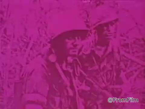 The still from the 1968 PSA 'Mail Early to Your Soldier' features two servicemen in military attire, likely captured in a jungle or dense foliage environment, indicative of the Vietnam setting. The color palette appears to be overwhelmingly pink, which may reflect a stylistic choice or a result of the film's aging. Both men seem serious as they stand together, suggesting camaraderie amidst challenging circumstances. The message reinforces the importance of sending mail and gifts to soldiers during the holiday season, encouraging timely preparation and clear addressing for packages to ensure they reach their recipients on time.