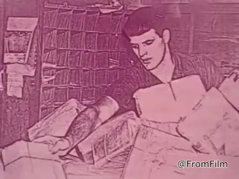 The still from the PSA 'Mail Early to Your Soldier' features a young serviceman in a postal setting. He appears to be sorting through packages, focusing on his task with a serious expression. The environment is cluttered with various mail items, likely related to holiday packages being prepared for shipping to soldiers overseas.   The visual tone is likely muted or stylized, reflecting the era of the 1960s, with a strong emphasis on the importance of timely mailing. The message of ensuring gifts reach servicemen before the deadline of November 9th suggests a sense of urgency and care, aimed at fostering connection and support for those serving in Vietnam during the holiday season.