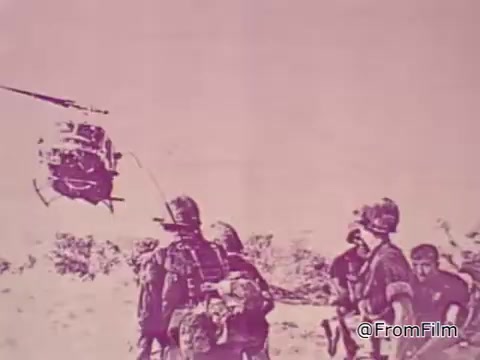 The still from the 1968 PSA 'Mail Early to Your Soldier' depicts a group of soldiers in a seemingly barren landscape, likely in Vietnam. In the background, a helicopter can be seen hovering, creating a sense of urgency and action. The soldiers are dressed in military gear, some with helmets and equipment, suggesting they are engaged in or preparing for active duty. The entire image has a pinkish tint, which may reflect the era's film quality or a stylistic choice to convey a specific mood. The voice-over emphasizes the importance of sending gifts to servicemen by a specific date to ensure they receive them in time for Christmas, underscoring a connection between families back home and their loved ones overseas.