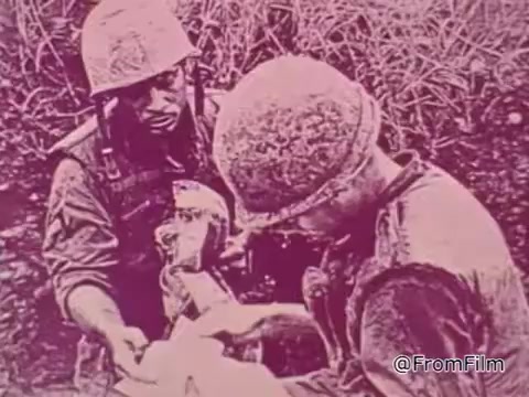 The still from the 1968 Vietnam PSA 'Mail Early to Your Soldier' captures a poignant moment between two servicemen. One soldier appears to be handing something to another, possibly a letter or package, emphasizing the importance of communication and connection during wartime. The soldiers are dressed in military attire, complete with helmets, and are surrounded by dense vegetation, indicative of the Vietnam landscape. The tone of the image is serious, reflecting the gravity of life in combat and the emotional weight of sending and receiving mail. The overall pinkish hue adds a nostalgic quality, resonating with the era's style and the sentiment of the message that encourages timely mailing before the holiday season.