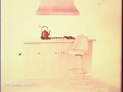The still features a minimalist kitchen scene, showcasing a simple cabinetry design and a stovetop with a kettle. A high-backed stool is positioned nearby, enhancing the domestic atmosphere. The color palette appears faded, lending a vintage quality to the image, typical of a 16mm film. The overall composition emphasizes functionality and cleanliness, in line with the commercial's purpose.