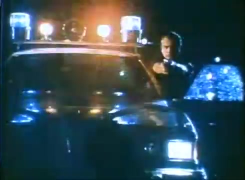 The still from the 'Love Child' trailer features a nighttime scene with a police car, its lights flashing. A figure stands near the car, holding a firearm, suggesting a tense or dramatic situation. The lighting and color palette reflect a moody atmosphere typical of suspenseful moments in film. The overall composition hints at potential conflict or action.
