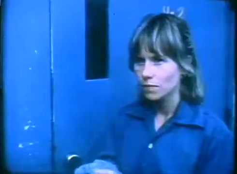 The still depicts a young woman, Amy Madigan's character, standing outside a blue door with a small window. She has shoulder-length hair and a serious expression, wearing a blue shirt. The lighting is somewhat muted, characteristic of 16mm film, creating a nostalgic, gritty atmosphere. The scene suggests tension or anticipation, fitting within the context of the 'Love Child' trailer.