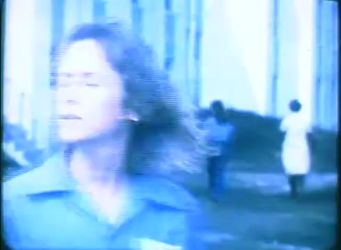 The still from the 'Love Child' trailer features a woman with slightly tousled hair, wearing a blue shirt. She appears to be in motion, looking away from the camera with an anxious expression. In the background, two other figures can be seen, adding a sense of context and urgency to the scene. The overall tone is muted, typical of a 16mm print, contributing to a nostalgic or tense atmosphere.