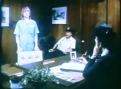 The still features a tense scene inside an office. A woman stands, dressed in a blue jumpsuit, looking apprehensive. Seated nearby is a man in uniform, observing her. At the desk, another woman is engaged in conversation, resting her chin on her hand, indicating intrigue or contemplation. The walls are adorned with framed pictures, contributing to the office atmosphere. The overall tone suggests a serious or dramatic moment in the film 'Love Child.'