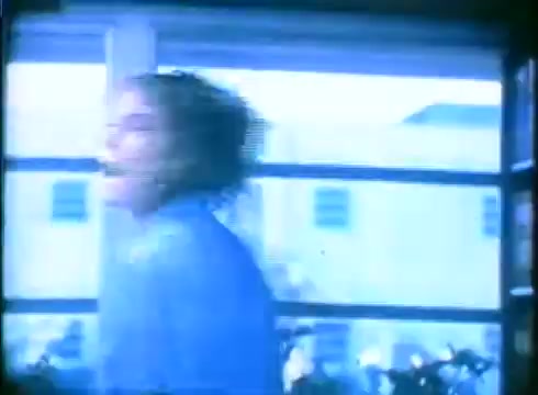 The still from the 'Love Child' trailer features a blurred, dynamic image of a woman, likely Amy Madigan, in motion. The scene appears to be shot indoors, with a bright, somewhat hazy ambiance from the natural light coming through a window. The background hints at an exterior view, while the focus on the woman's movement conveys a sense of urgency or emotional intensity. The color tones lean towards a cooler palette, typical of 16mm film aesthetics.