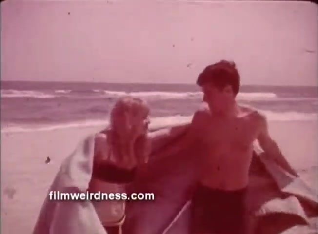The still is from a 1960s Nationwide Insurance commercial, featuring a young couple on a beach. The woman, dressed in a bikini, and the shirtless man are sharing a blanket, conveying a warm and relaxed atmosphere. The ocean is visible in the background, reflecting the carefree spirit of the era. The image has a vintage quality indicative of the film's age.