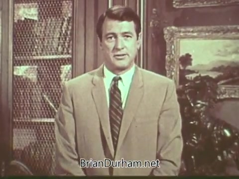 The still features a well-groomed man in a light-colored suit standing in front of a bookshelf. He appears serious and engaged, likely addressing the audience directly about multiple sclerosis. The background suggests a formal setting, with decorative elements that enhance the gravity of the message. This PSA aims to raise awareness about multiple sclerosis, highlighting its impact and the importance of understanding the condition.