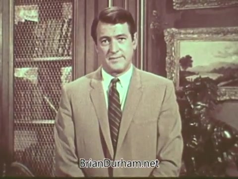 The still from the 'Rock Hudson Multiple Sclerosis PSA' features Rock Hudson, dressed in a light-colored suit and tie, standing against a backdrop of bookshelves and framed artwork. He appears serious and engaged, likely delivering a message about multiple sclerosis awareness. The image captures a moment from the public service announcement aimed at educating viewers on the condition.