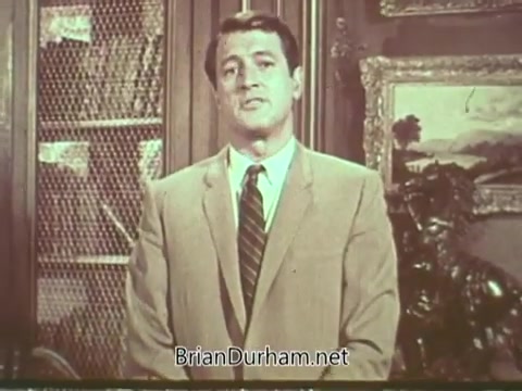 The still from the 'Rock Hudson Multiple Sclerosis PSA' features Rock Hudson standing in a formal setting. He is dressed in a light-colored suit with a tie, addressing the audience. The background includes shelves, possibly containing books, and a painted landscape scene. The overall atmosphere conveys a sense of seriousness and concern regarding multiple sclerosis.