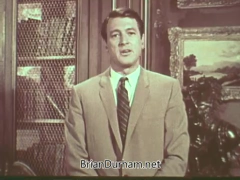 The still from the 'Rock Hudson Multiple Sclerosis PSA' features Rock Hudson, who is dressed in a light suit and tie, standing in an indoor setting. Behind him is a backdrop that includes framed artwork and a bookshelf. Hudson appears earnest and engaging, likely addressing the audience with a message about multiple sclerosis, a condition he was known to have publicly diagnosed. The visual reflects a classic mid-20th century television style.