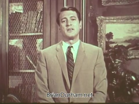 The still features Rock Hudson, dressed in a light-colored suit and tie, standing in front of a decorative backdrop that includes bookshelves and a painting. He appears earnest as he addresses the audience, likely discussing the topic of multiple sclerosis in a public service announcement. The overall tone is informative and supportive, aimed at raising awareness about the condition.