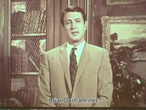 The still features Rock Hudson standing in a well-decorated room, dressed in a light-colored suit with a tie. He appears to be addressing the audience directly, likely conveying an important message about multiple sclerosis. The background includes shelves and decorations that suggest a warm, cultured environment, emphasizing the seriousness of the topic he is discussing.