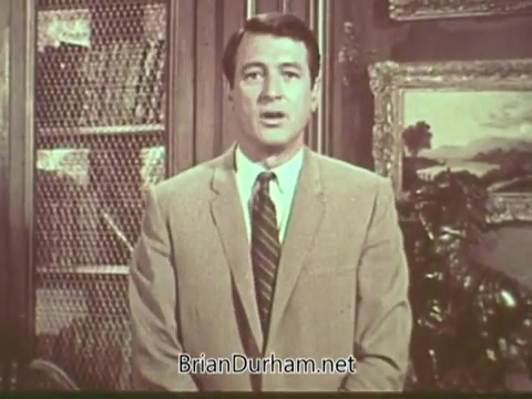 The still features Rock Hudson, dressed in a suit with a tie, addressing the audience. It appears to be a public service announcement (PSA) focused on multiple sclerosis. The background includes a bookshelf and a framed landscape painting, creating a formal setting. Hudson's expression is serious, reflecting the importance of the topic at hand.