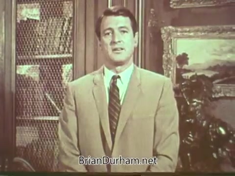 The still features Rock Hudson, dressed in a light-colored suit and tie, standing in front of a bookshelf and artwork. He appears earnest and concerned, delivering a public service announcement about multiple sclerosis. The background suggests a formal setting, contributing to the seriousness of the topic.