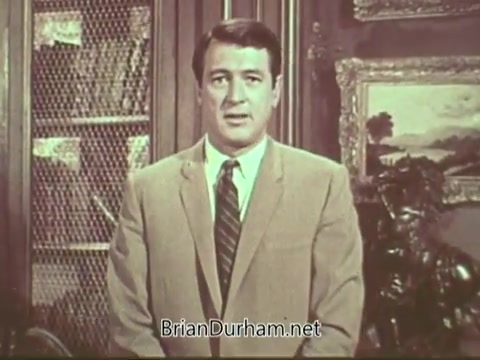 The still from the 'Rock Hudson Multiple Sclerosis PSA' features a man in a light suit and tie, standing in front of a bookcase. The background includes decorative art pieces, suggesting a formal setting. His expression appears serious as he addresses the camera, likely conveying an important message about multiple sclerosis awareness.