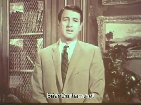 The still shows Rock Hudson, dressed in a light-colored suit with a tie, speaking directly to the camera. The background features bookshelves, decorative frames, and a statue, suggesting a formal setting. Hudson's serious expression indicates the importance of the message he is delivering about multiple sclerosis awareness. The overall tone is informative and earnest.