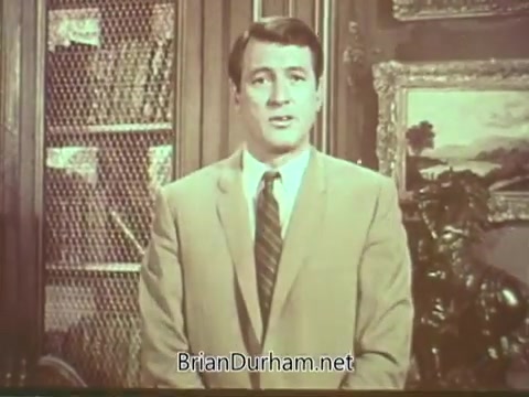The still shows Rock Hudson in a formal setting, wearing a light-colored suit and a tie, addressing the camera. The background features bookshelves and decorative elements, suggesting a classic or professional atmosphere. This PSA aims to raise awareness about multiple sclerosis.