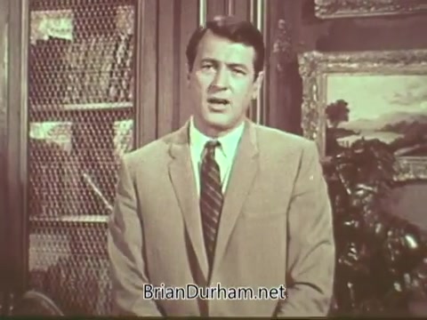 The still features Rock Hudson, dressed in a light-colored suit and tie, standing in a well-furnished, classic room. Behind him are shelves, likely filled with books, and decorative elements that suggest a sophisticated setting. Hudson's expression is serious as he addresses the camera, likely conveying an important message about multiple sclerosis. The image captures the earnest tone of the public service announcement.