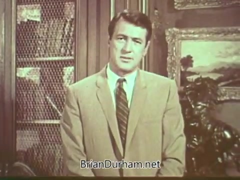 The still features Rock Hudson in a formal setting, wearing a light-colored suit and tie. He appears serious as he addresses the camera, likely raising awareness about multiple sclerosis. The background includes a bookshelf and framed artwork, contributing to a classic, sophisticated atmosphere. This PSA is significant as it highlights Hudson's role in increasing public awareness of the disease during a time when it was less understood.