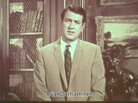 The still features Rock Hudson delivering a public service announcement about multiple sclerosis. He appears in a tailored suit, standing in front of a bookshelf and artwork. The setting conveys a sense of seriousness, reflecting the importance of the message regarding awareness and education about the disease. Hudson looks directly into the camera, engaging the audience with a sincere expression.