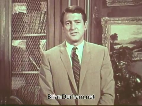 The still from the 'Rock Hudson Multiple Sclerosis PSA' features Rock Hudson delivering a message in a formal setting. He is dressed in a light-colored suit with a tie, standing in front of a background that includes bookshelves and decorative artwork. The mood appears serious yet hopeful, reflecting the informative nature of the public service announcement aimed at raising awareness about multiple sclerosis.